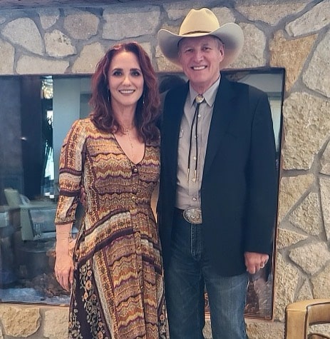 Bruce Boxleitner in black coat, blue jeans, and bone white color hat smiling with his wife in a brown dress.